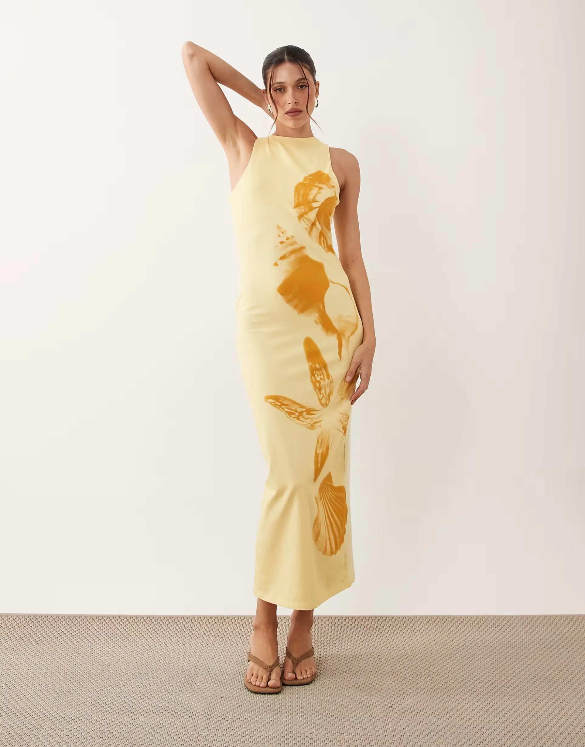 Model in a buttery-yellow ASOS DESIGN maxi dress with an abstract orange shell and floral print, worn with tan strappy sandals against a neutral studio backdrop.