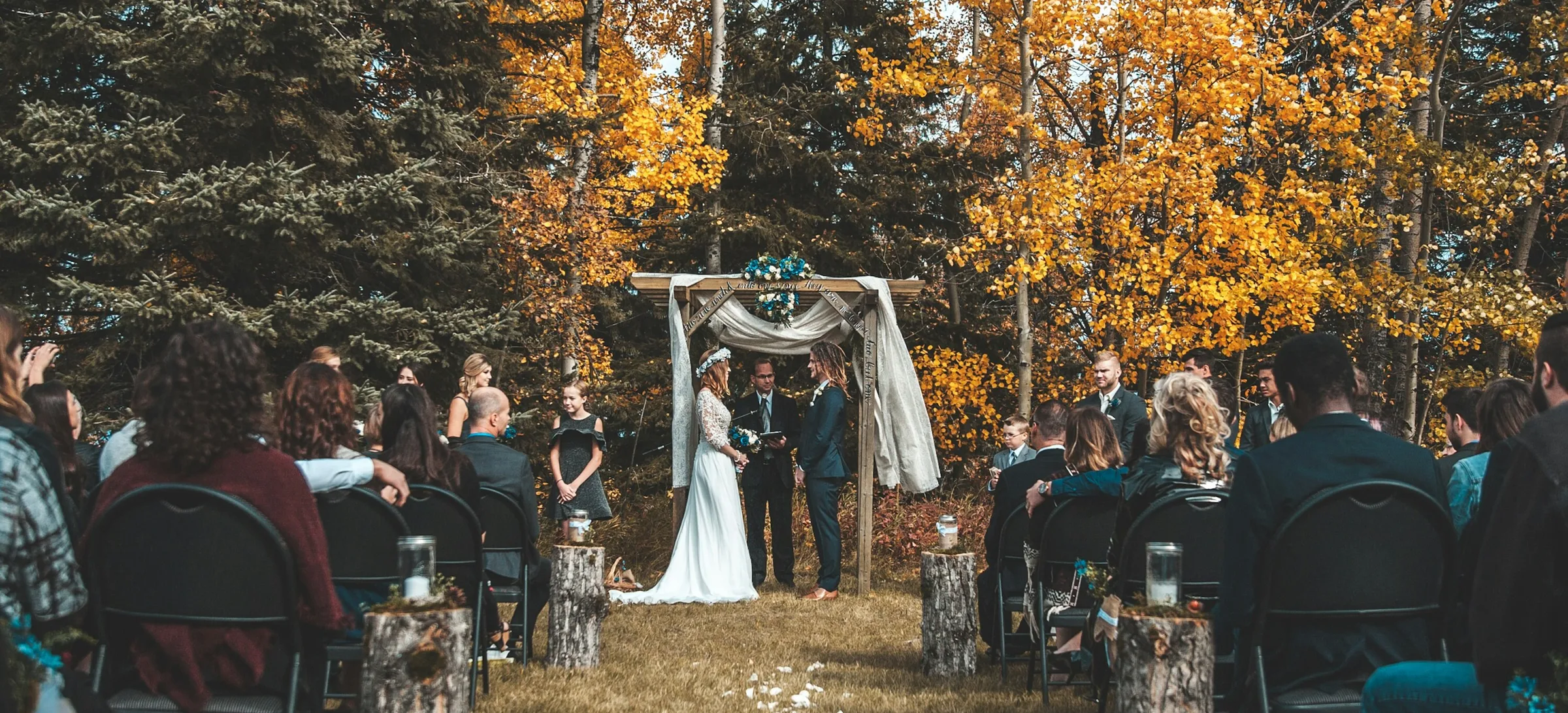 Guests seated on black chairs either side of a grass aisle at an outdoor autumn wedding ceremony, several wrapped in shawls and coats, with golden aspen and dark pine trees behind the couple and officiant under a draped wooden arch.