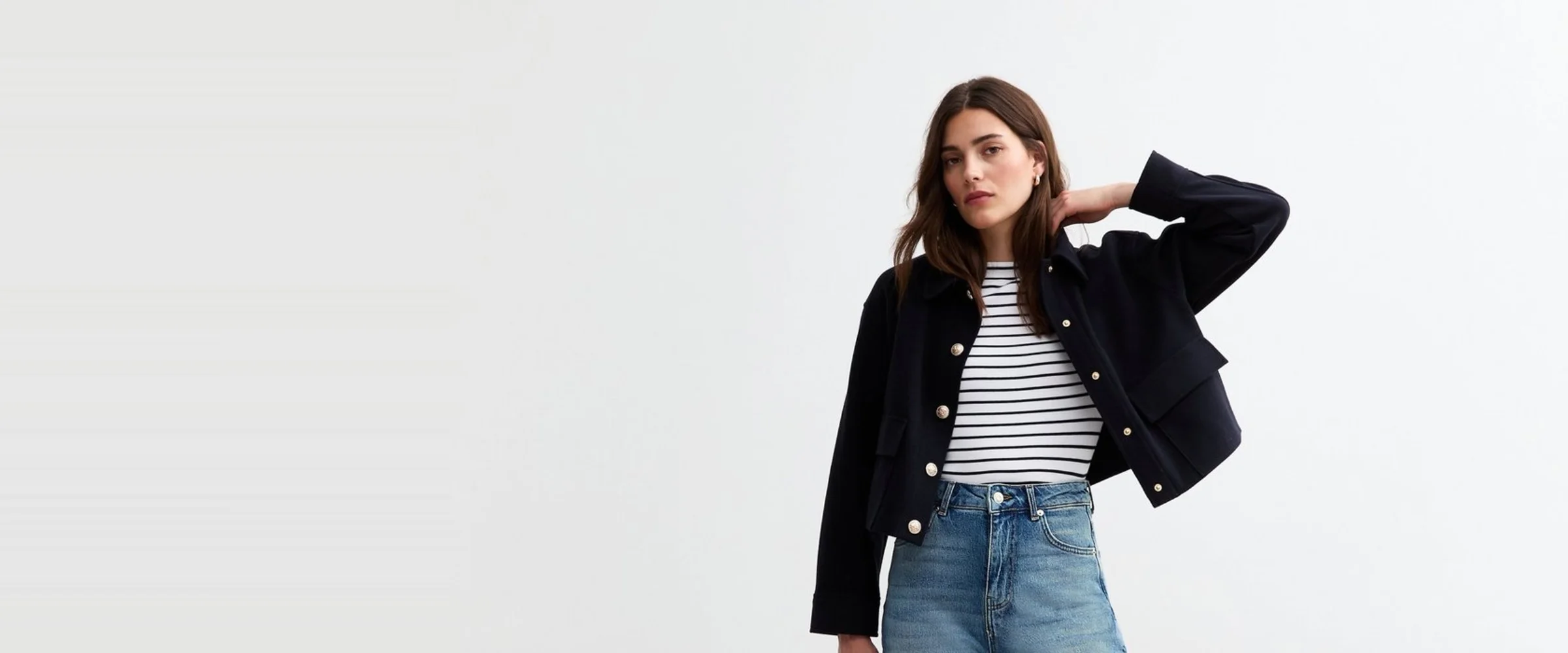 A model in high-waisted straight-leg blue jeans, a navy and white breton stripe top and a cropped navy jacket with gold buttons, holding a small black bag