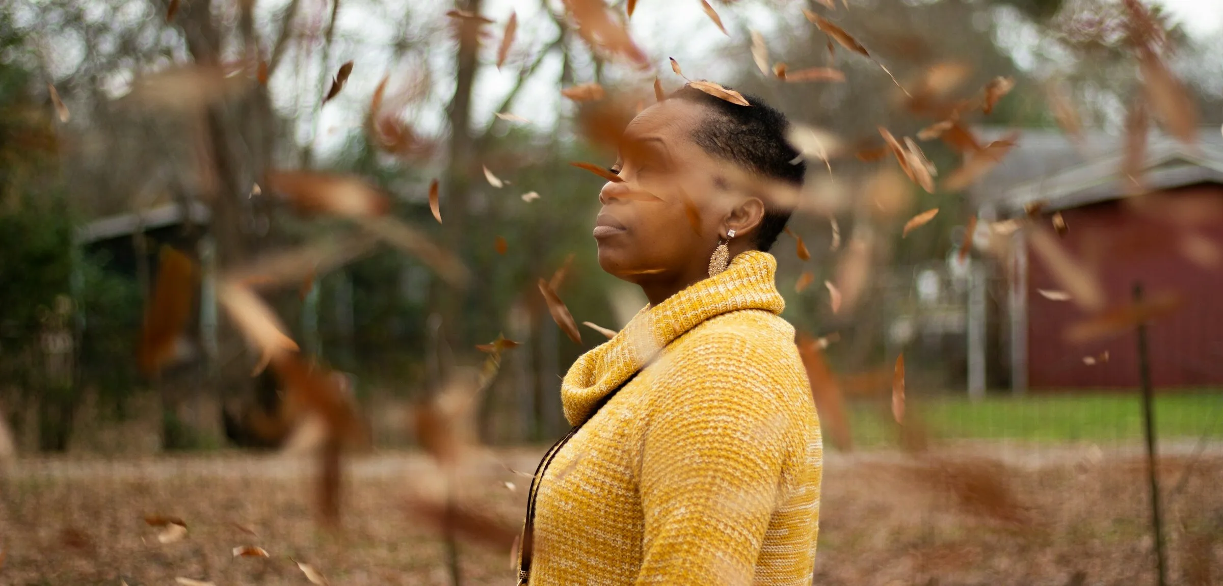 A woman in a mustard-yellow ribbed roll-neck knit standing outdoors as dry autumn leaves blow past her