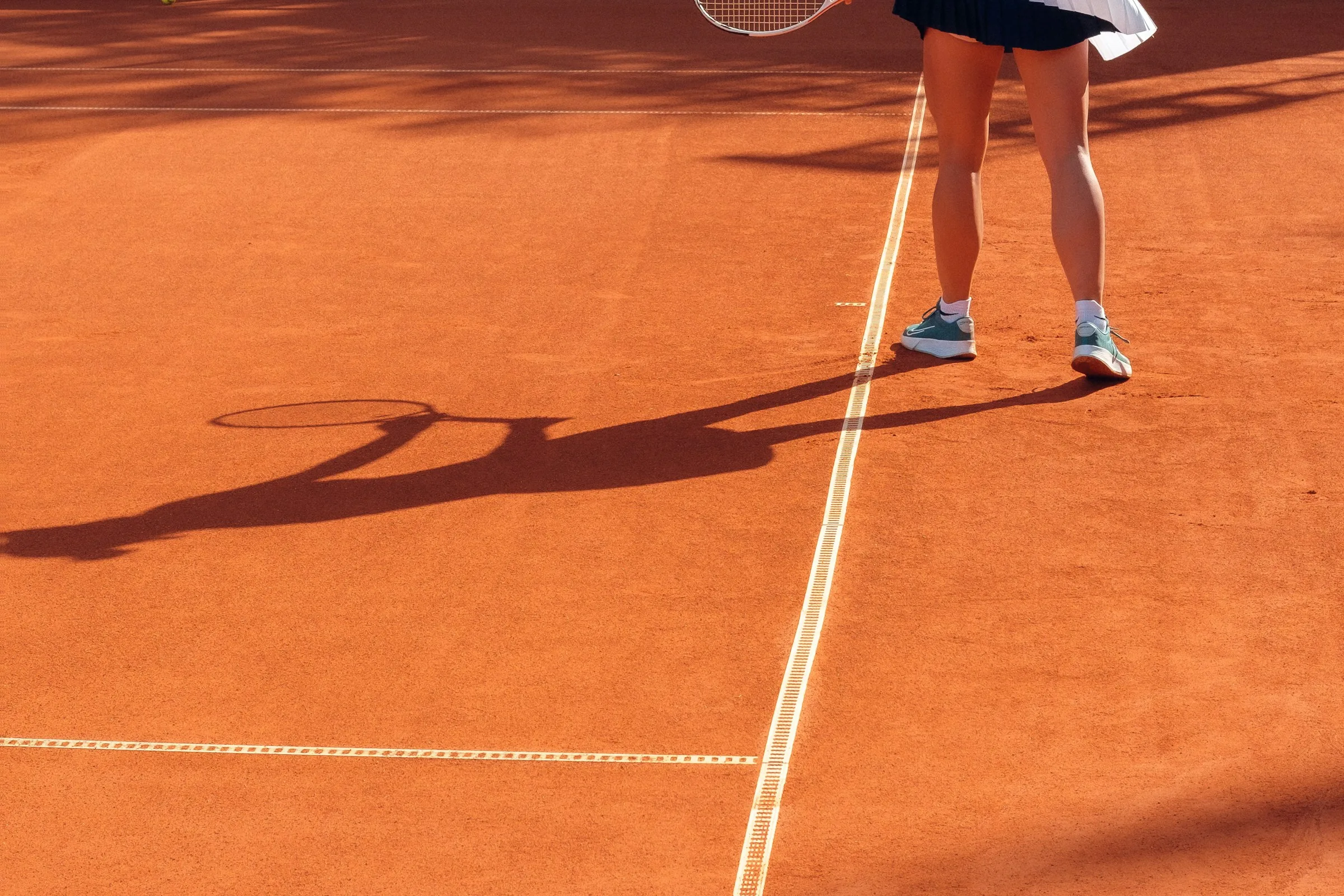 A tennis player in a navy pleated skirt and white trainers casts a long racket shadow across an orange clay court