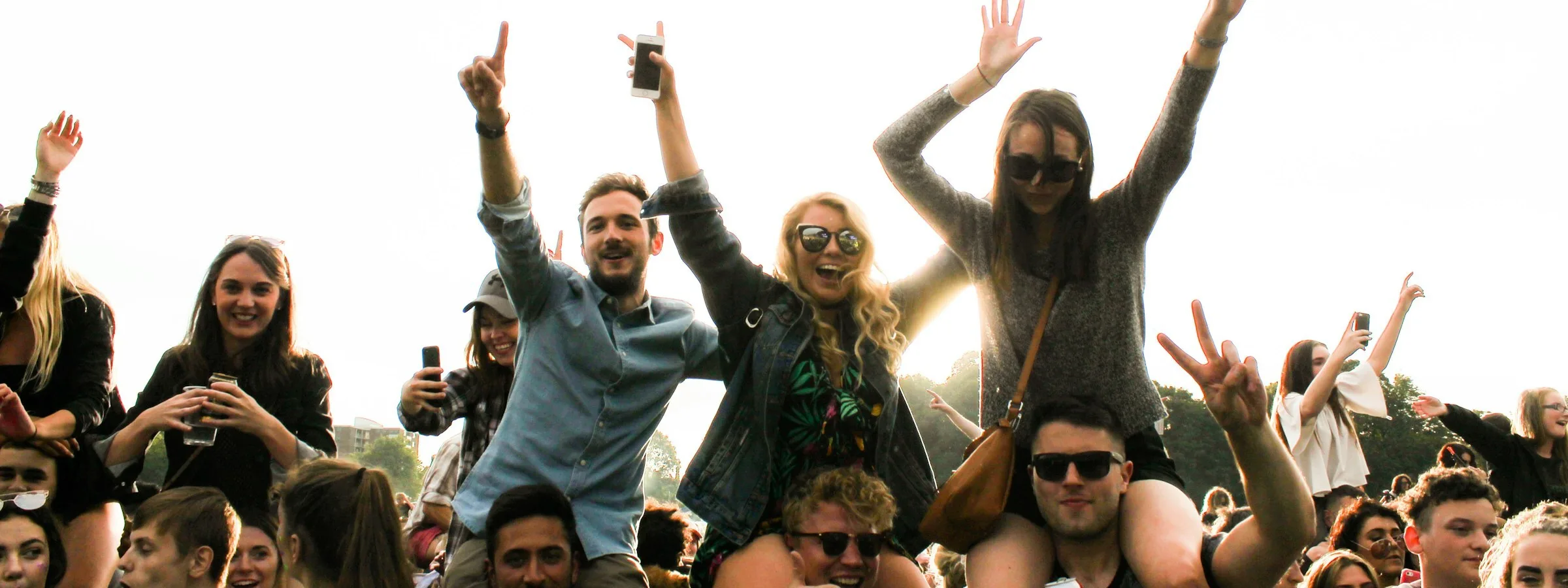 A crowd of young people at an outdoor festival in late afternoon sun, several sitting on shoulders with arms raised, wearing denim jackets and knitwear, cans in hand