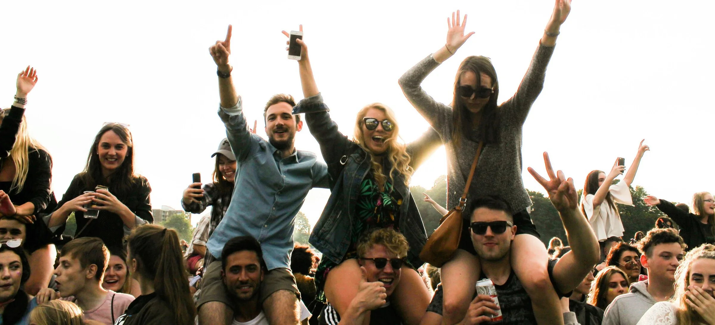 A crowd at an outdoor evening gig on grass in a UK park, backlit by low sun, with people up on shoulders in a denim jacket, a black jacket and a grey knit, hands in the air