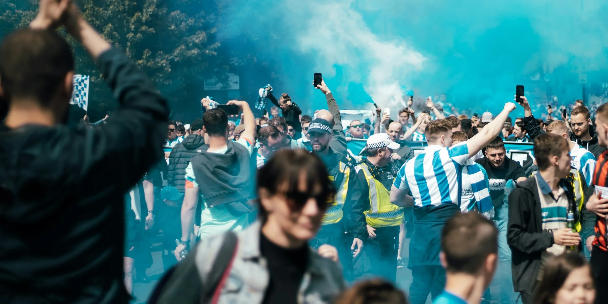 Football supporters walking to a match through pale blue smoke, in striped shirts, hoodies and jackets, with a police officer in a hi-vis jacket among the crowd