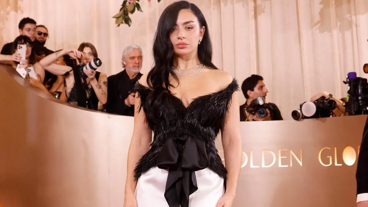 Charli XCX at the 2026 Golden Globes in an off-the-shoulder black feathered Saint Laurent bodice tied with a black satin bow over a white column skirt, with layered diamond necklaces and photographers behind her