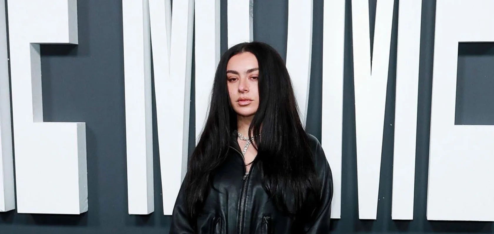 Charli XCX in a zipped black leather jacket and a diamond necklace in front of giant white letters spelling The Moment at the film's London premiere