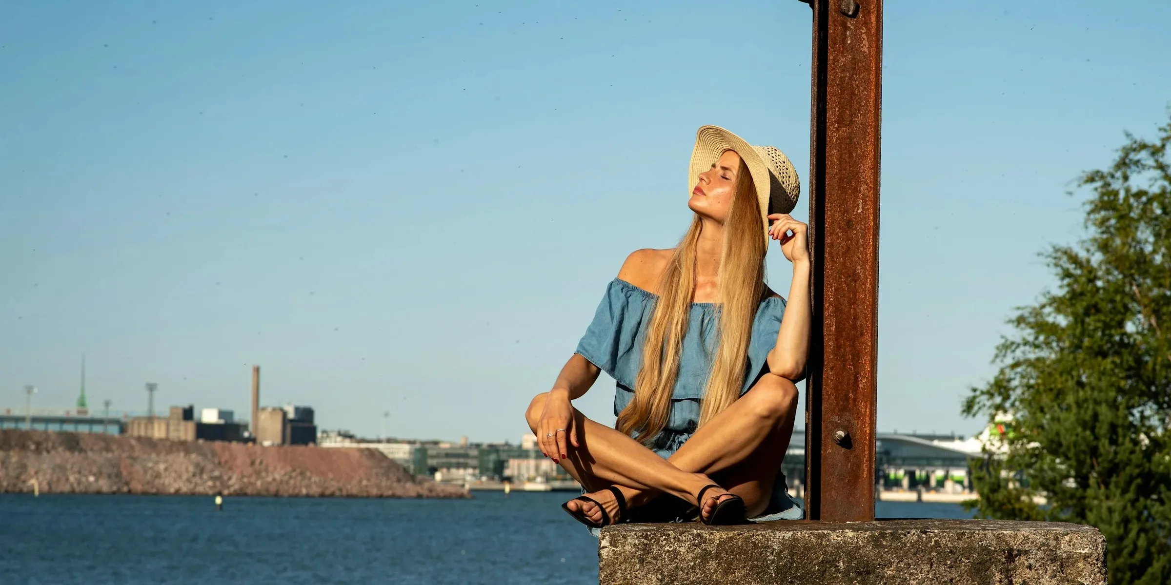 Woman in a straw hat and chambray off-shoulder top sitting on a harbourside wall in golden evening light, sea and dockyard skyline behind her