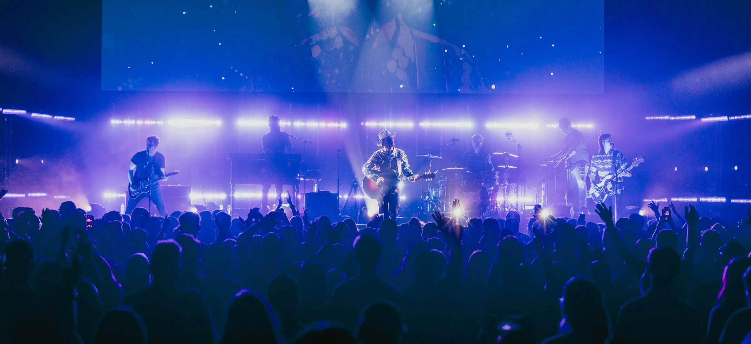 A six-piece guitar band on a wide arena stage under purple and white lights, with a packed standing crowd in silhouette in the foreground, some with hands raised.
