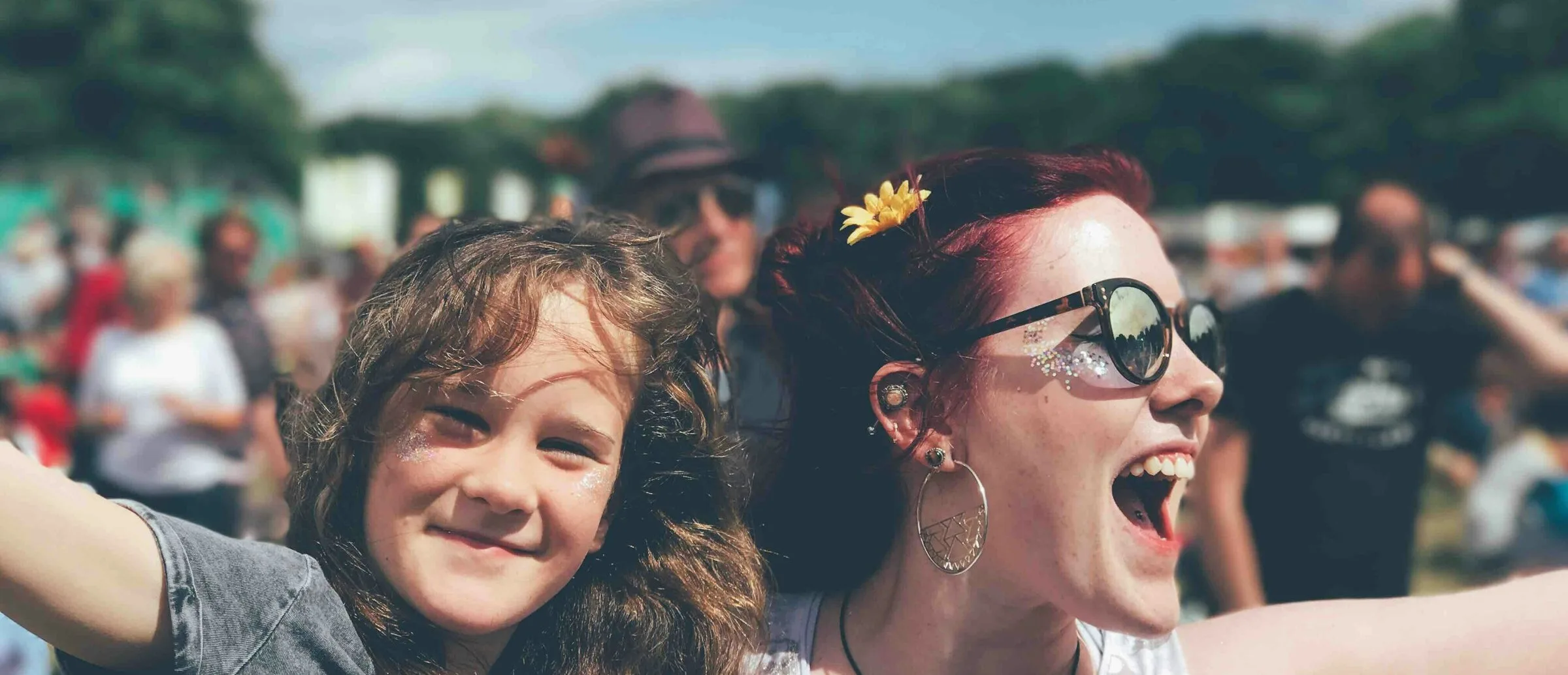 A woman in a white vest top with glitter under her eyes, hoop earrings, sunglasses and a yellow flower in her hair, arms outstretched beside a young girl in a grey graphic tee, both laughing in a sunny daytime festival crowd on grass