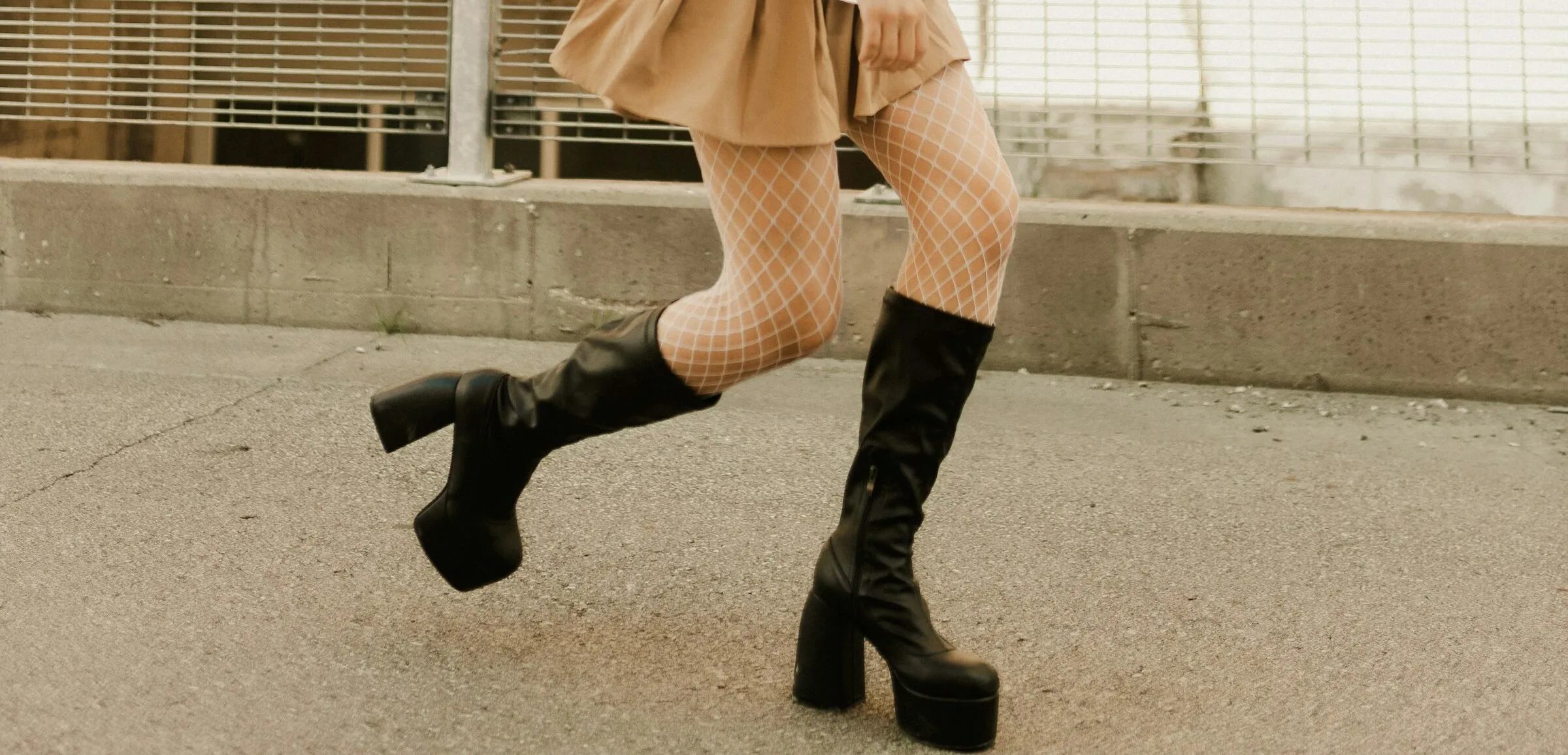 A woman mid-stride on a city street in black knee-high platform boots, a tan pleated skirt and white fishnet tights