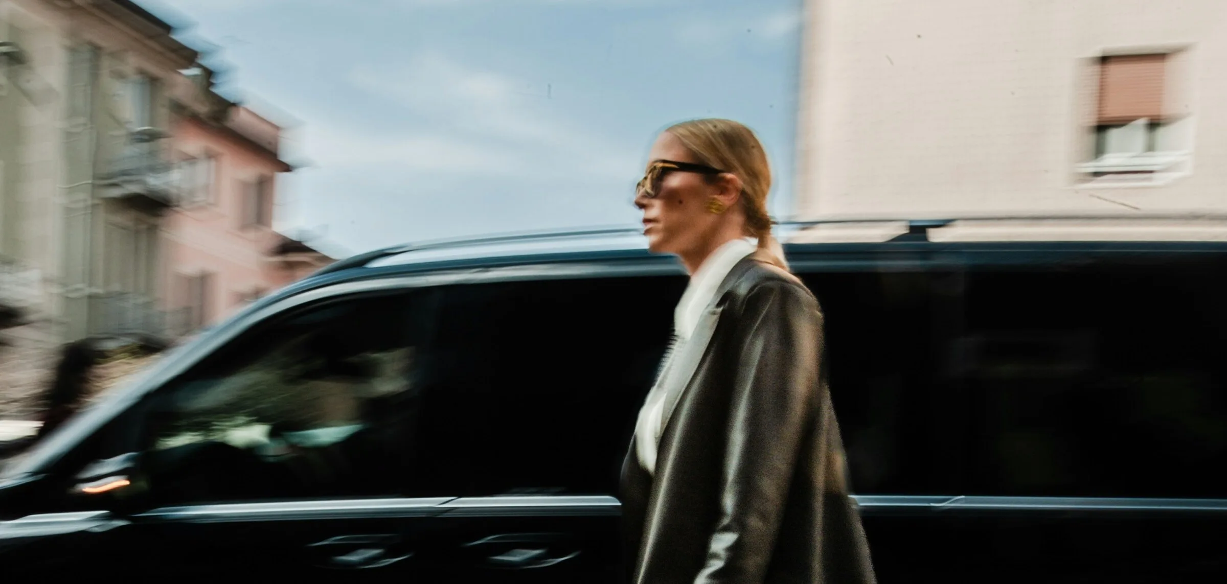 A woman in dark sunglasses and a long dark leather coat walking past a blurred black van on a city street, carrying a small gold top-handle bag.