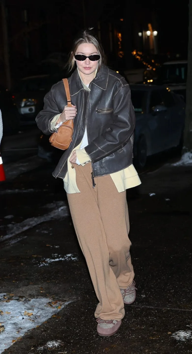 Gigi Hadid at night in New York in an oversized dark brown leather jacket over a butter-yellow cashmere jacket and camel trousers, with dark sunglasses and a tan shoulder bag