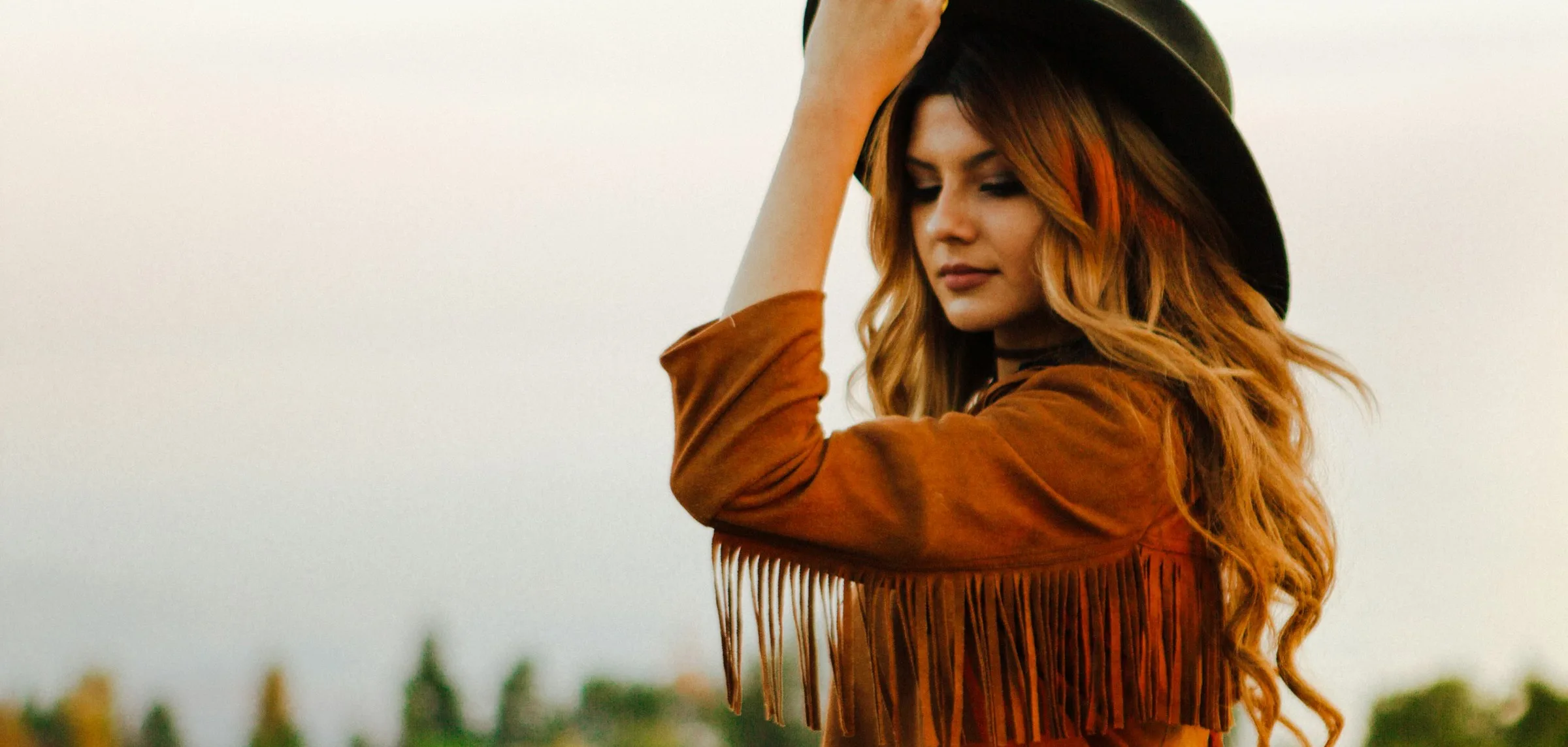 A woman in a cognac suede jacket with long fringe across the back, one hand holding the brim of a dark wide-brimmed felt hat, standing against a pale sky with autumn trees behind her.