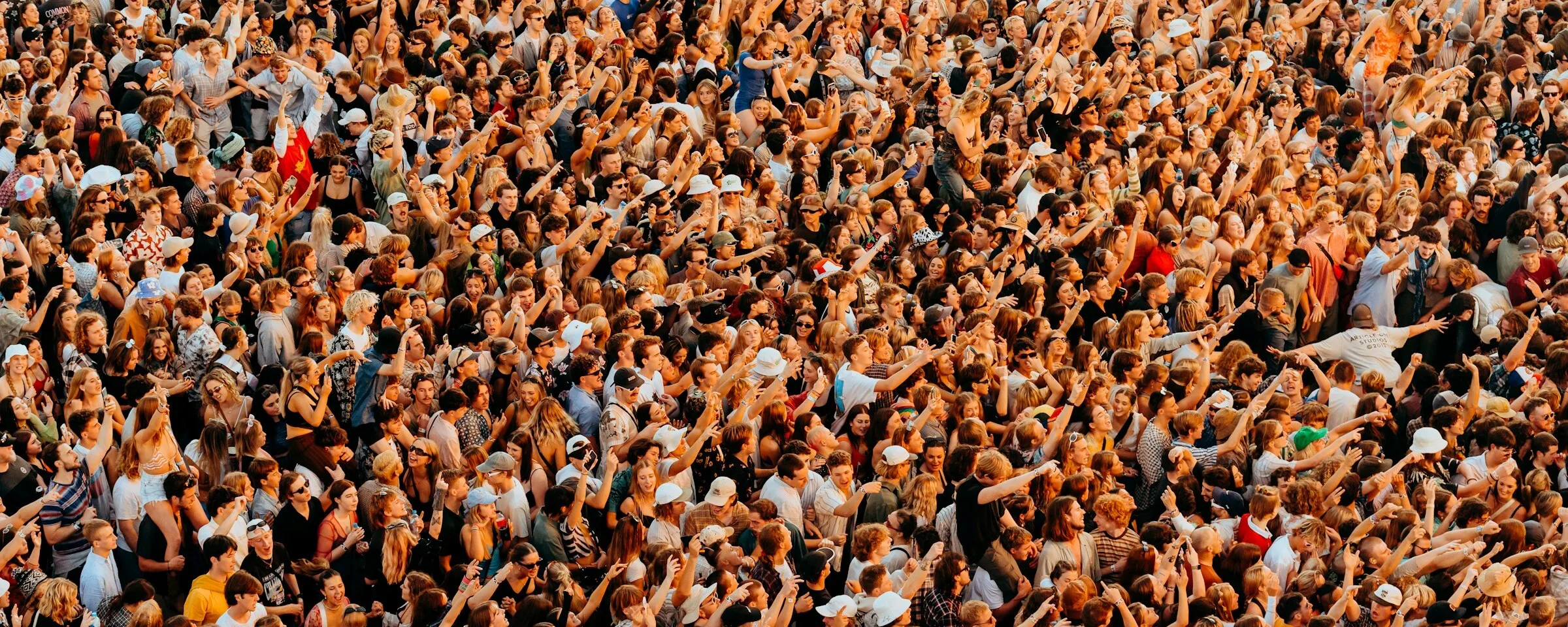Aerial view of a packed open-air gig crowd in golden evening light, arms raised, in vests, caps and sunglasses