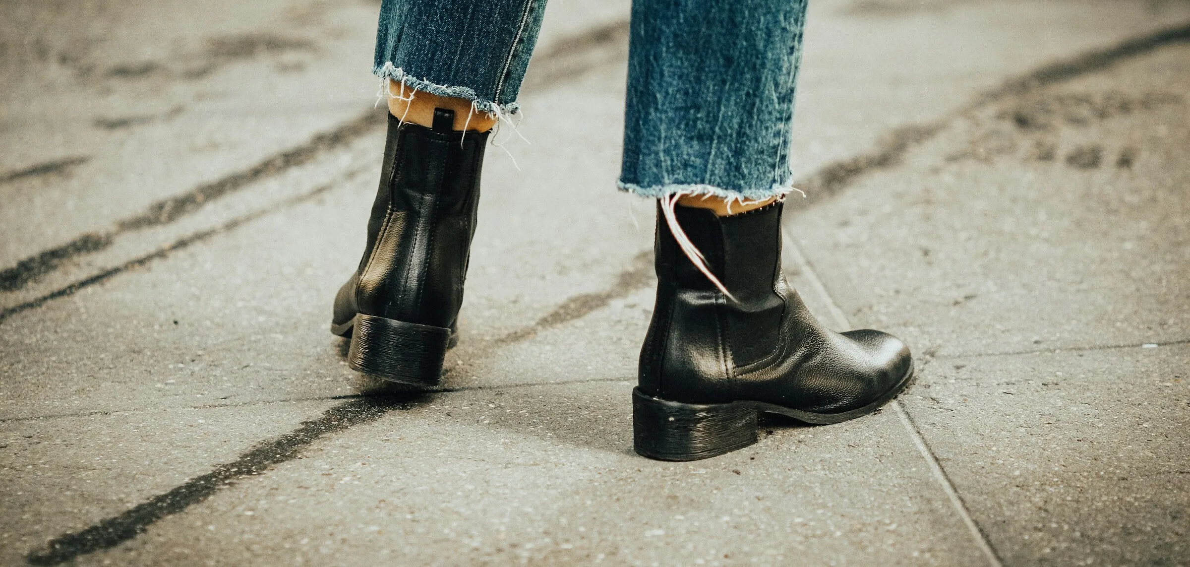 Close-up of black leather Chelsea-style ankle boots worn with raw-hem straight-leg blue jeans on a city pavement