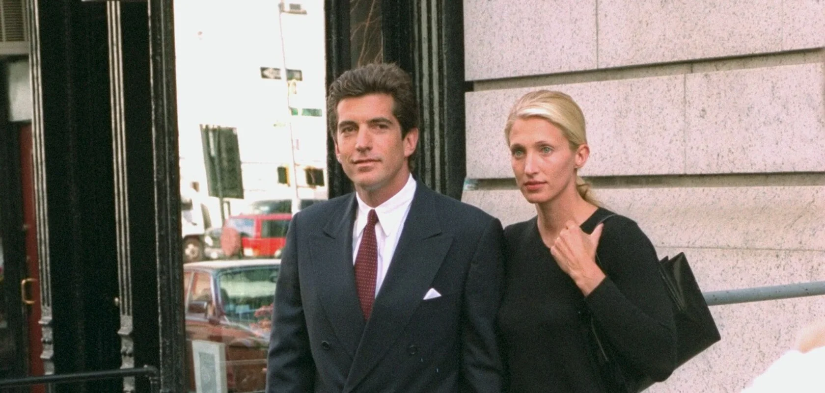 John F. Kennedy Jr. in a navy double-breasted suit and Carolyn Bessette-Kennedy in a black knit with a black shoulder bag, outside their Tribeca building in October 1996