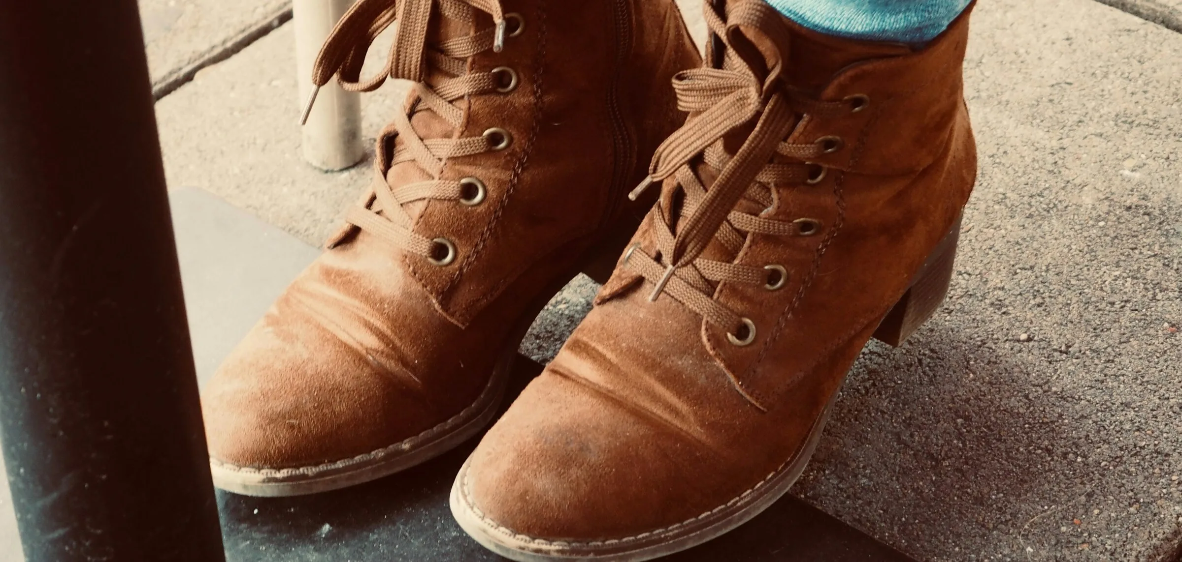 Close view of a pair of tan suede lace-up ankle boots with a low block heel, worn with light blue jeans on a pavement.