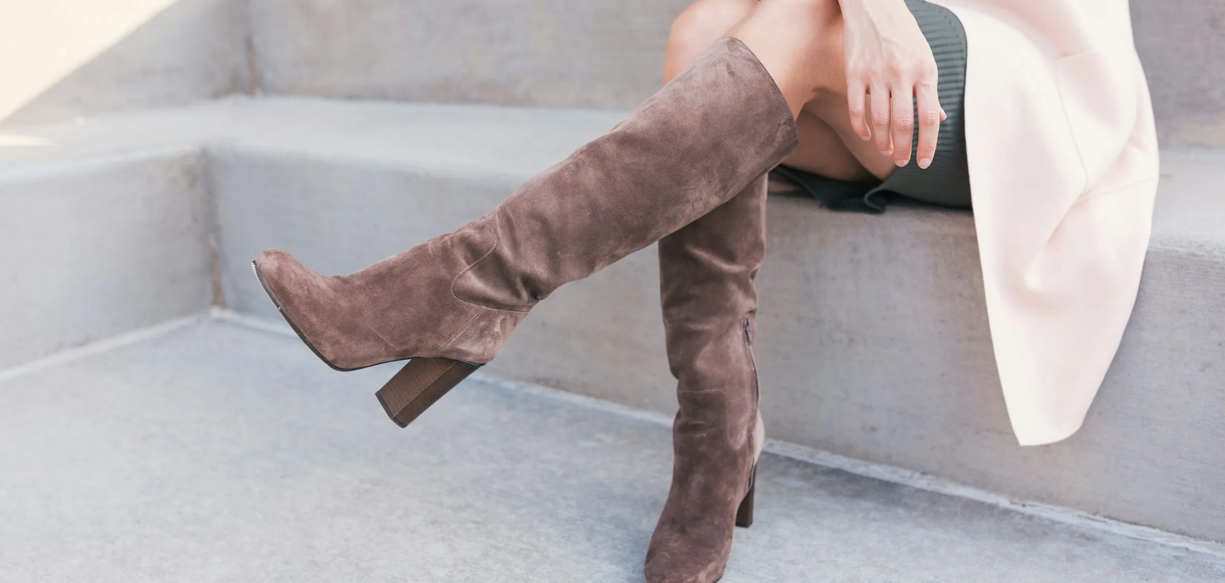 A woman sitting on concrete steps in taupe suede knee-high boots with a block heel, wearing a pale pink coat.