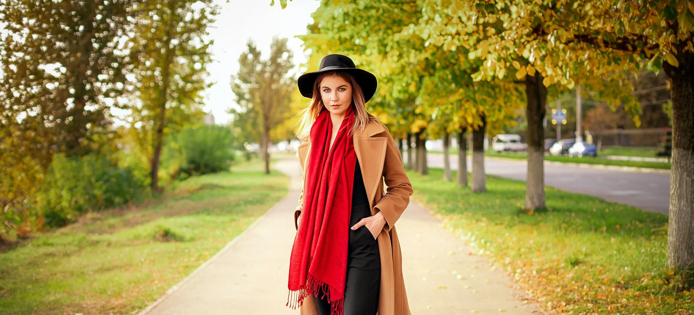 Woman in a long camel wool coat, black wide-brimmed hat and red scarf walking along a tree-lined path in autumn