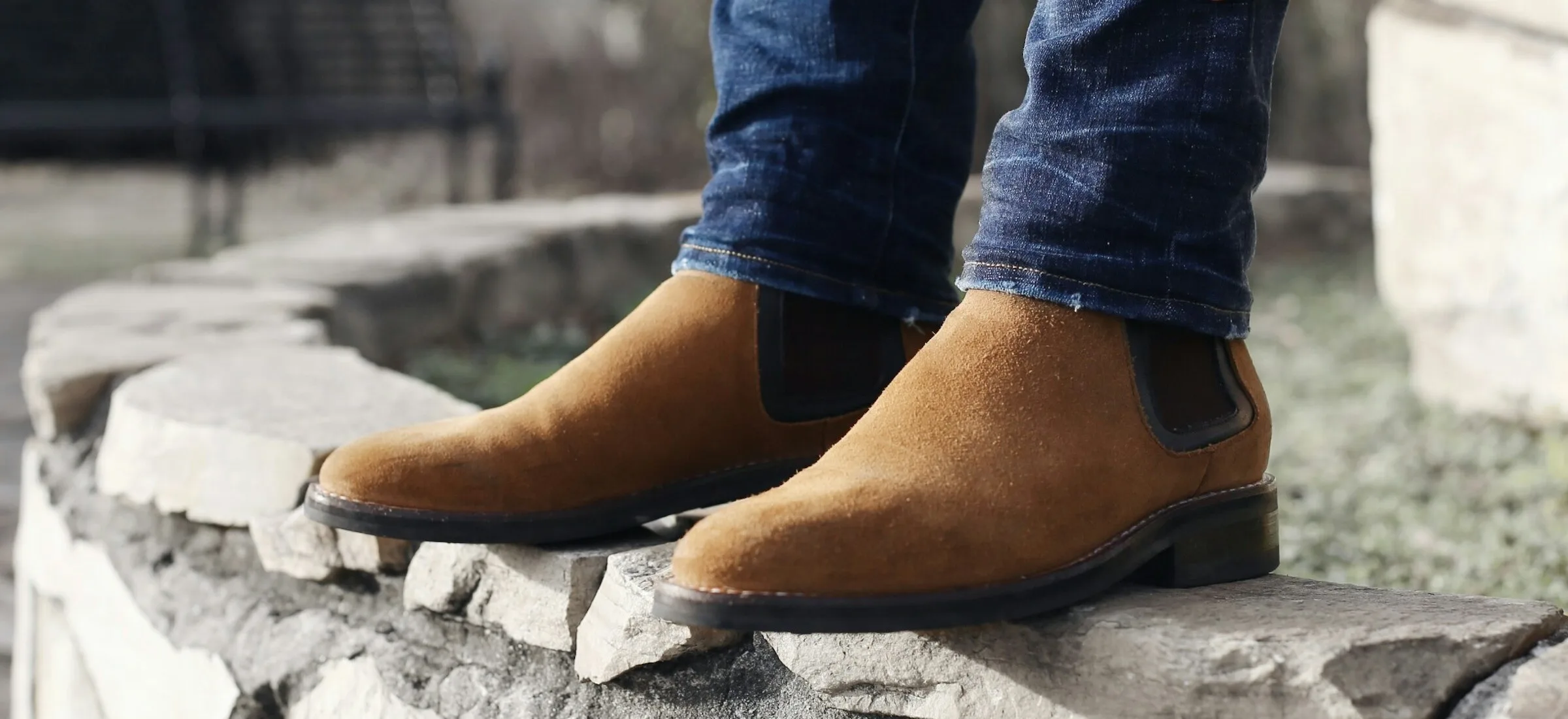 Close-up of a man in tan suede Chelsea boots and turned-up dark blue jeans, standing on a low stone wall outdoors.