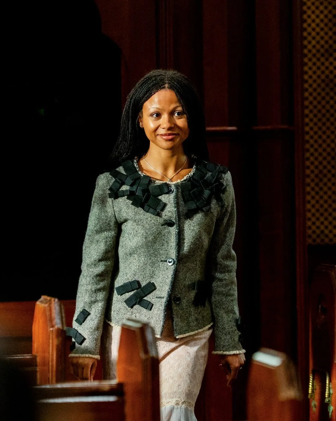 Myha'la walking through the castle chapel in a grey tweed jacket covered in small black ribbon bows, worn with a pale pink lace skirt