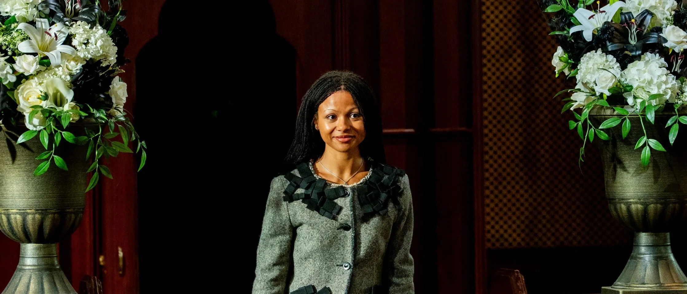 Myha'la smiling as she walks between pews and white flower arrangements in the castle chapel, wearing a grey tweed jacket covered in black ribbon bows