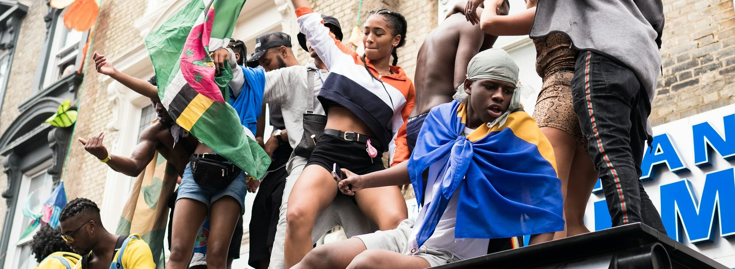 Revellers dancing on a bus shelter roof at Notting Hill Carnival, draped in Dominican and Barbadian flags, in crop tops, denim shorts and tracksuits