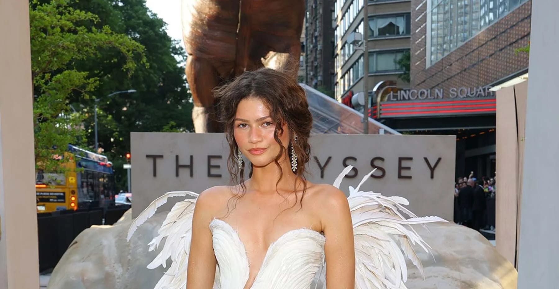 Zendaya on the red carpet at The Odyssey New York premiere in a strapless ivory pleated gown with feathered wings, in front of the film's stone signage at Lincoln Square.