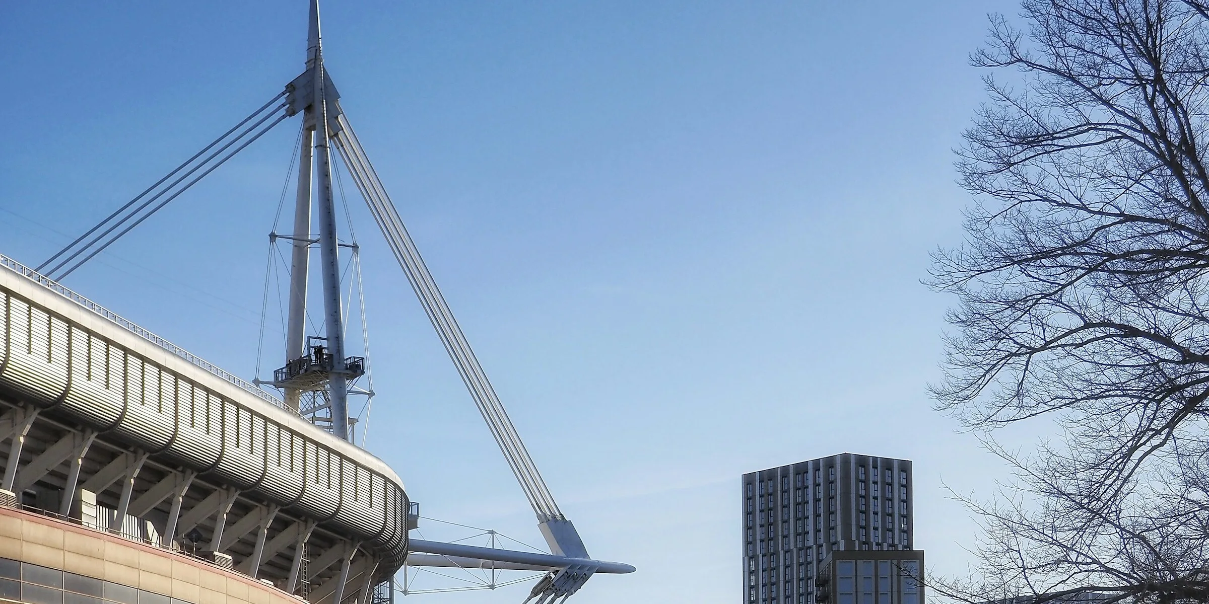 The Principality Stadium's mast and curved roof rising above Cardiff city centre against a clear blue sky, with a residential tower and bare winter trees to the right.