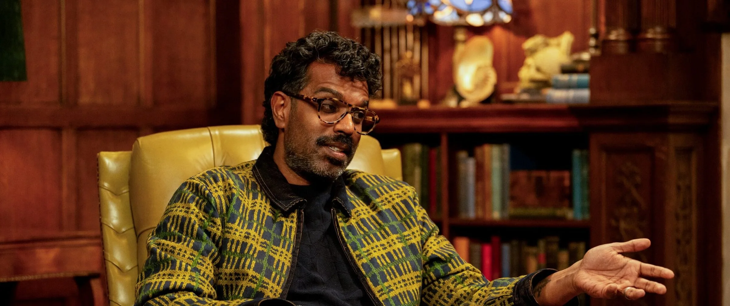 Romesh Ranganathan in a leather wingback chair in a wood-panelled Celebrity Traitors room, wearing a yellow, green and navy check knitted zip jacket over a navy knit, with tortoiseshell glasses and black beaded bracelets.