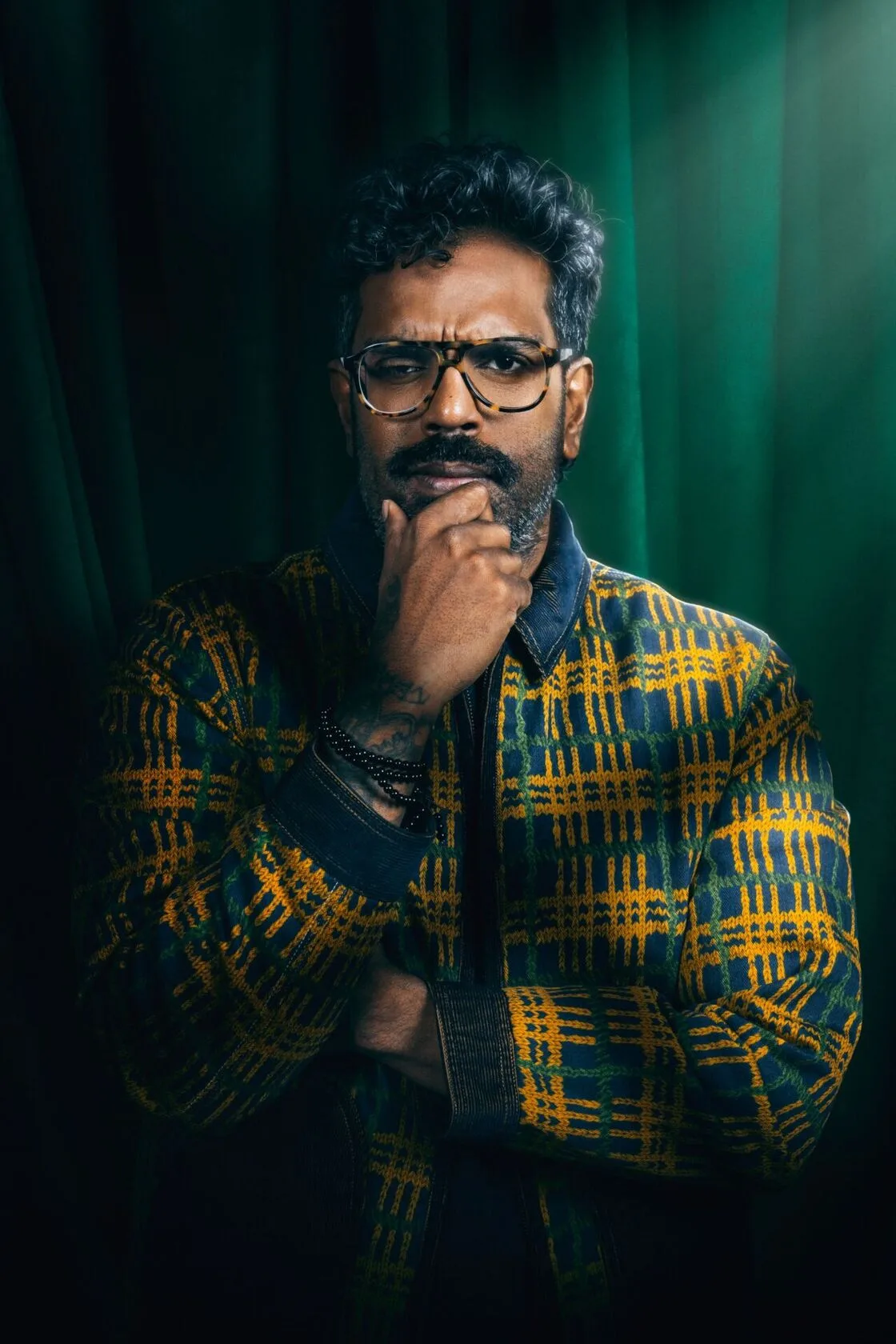 Romesh Ranganathan in his Celebrity Traitors cast portrait against green velvet curtains, hand on his chin, wearing a mustard, green and navy check knitted zip jacket with a dark collar, tortoiseshell glasses and black beaded bracelets.