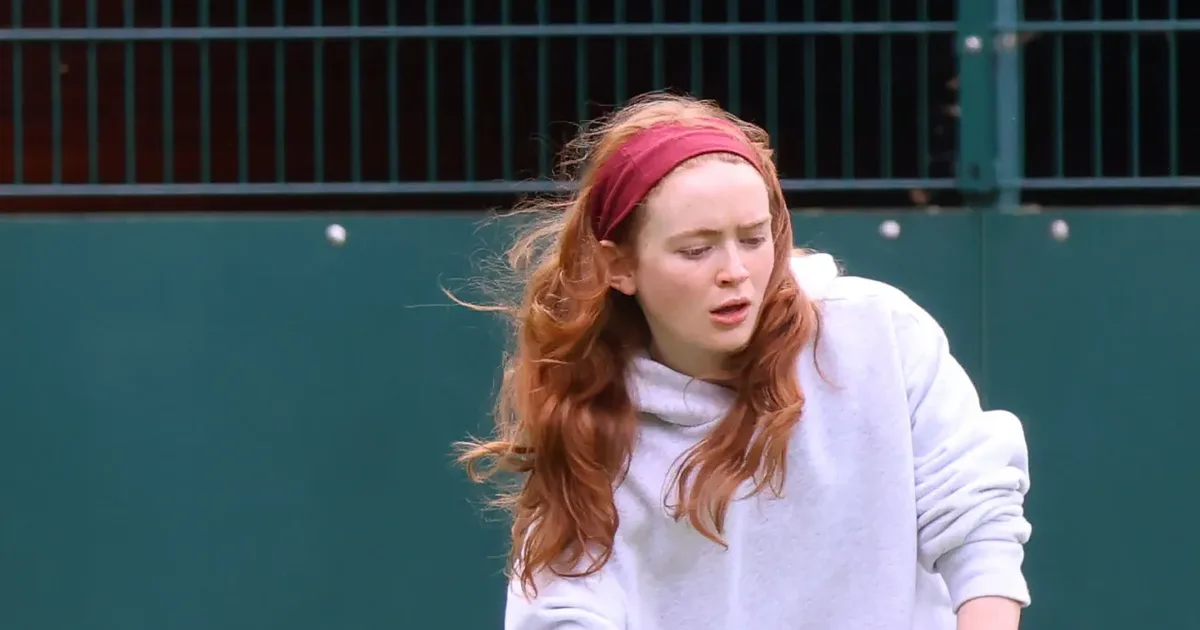Sadie Sink mid-serve at a tennis lesson in London, wearing an oversized grey hoodie, black leggings and a burgundy headband.