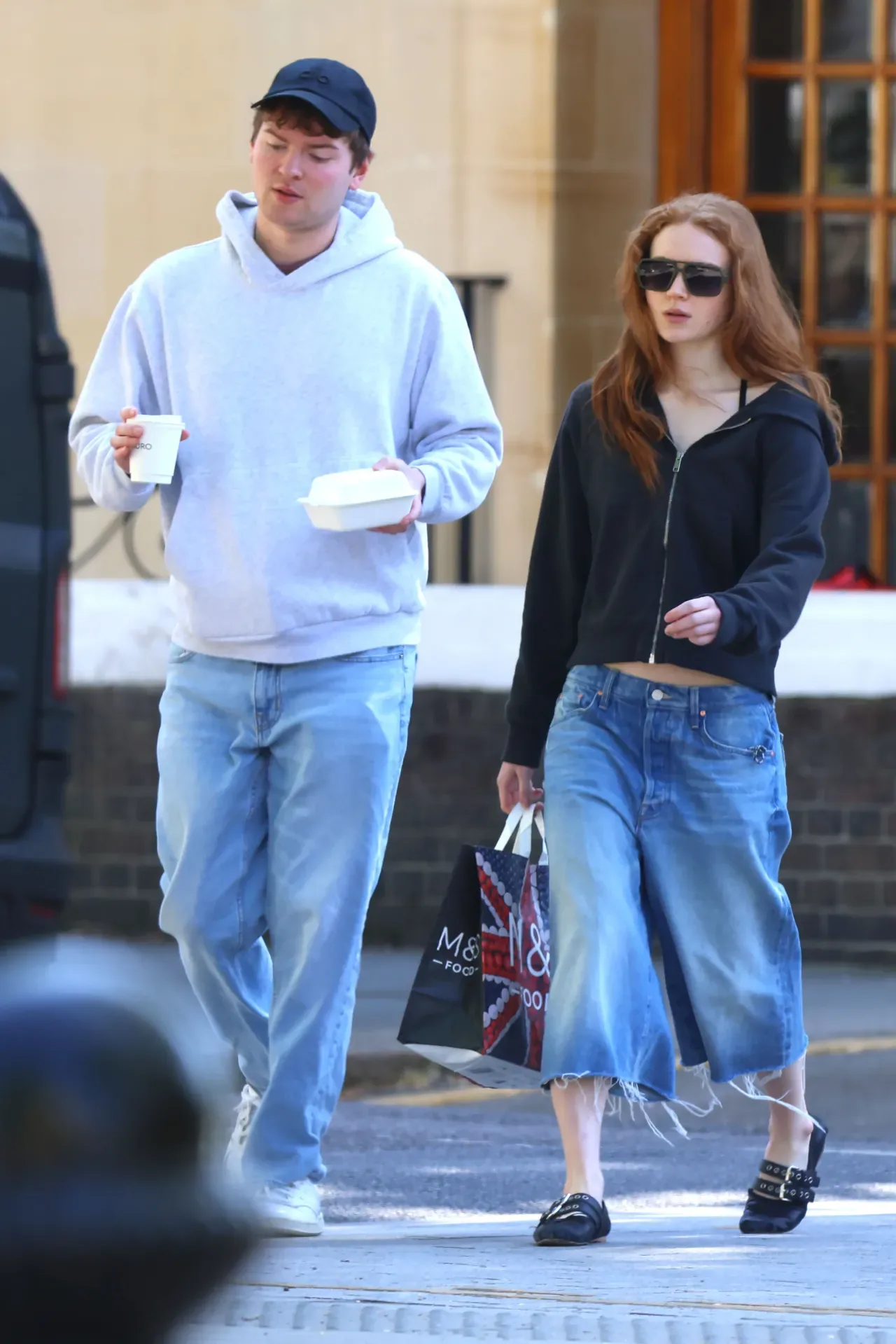 Sadie Sink walking in Notting Hill, London, wearing a cropped black zip hoodie, a wide raw-hem denim skirt, black flat mules and aviator sunglasses.