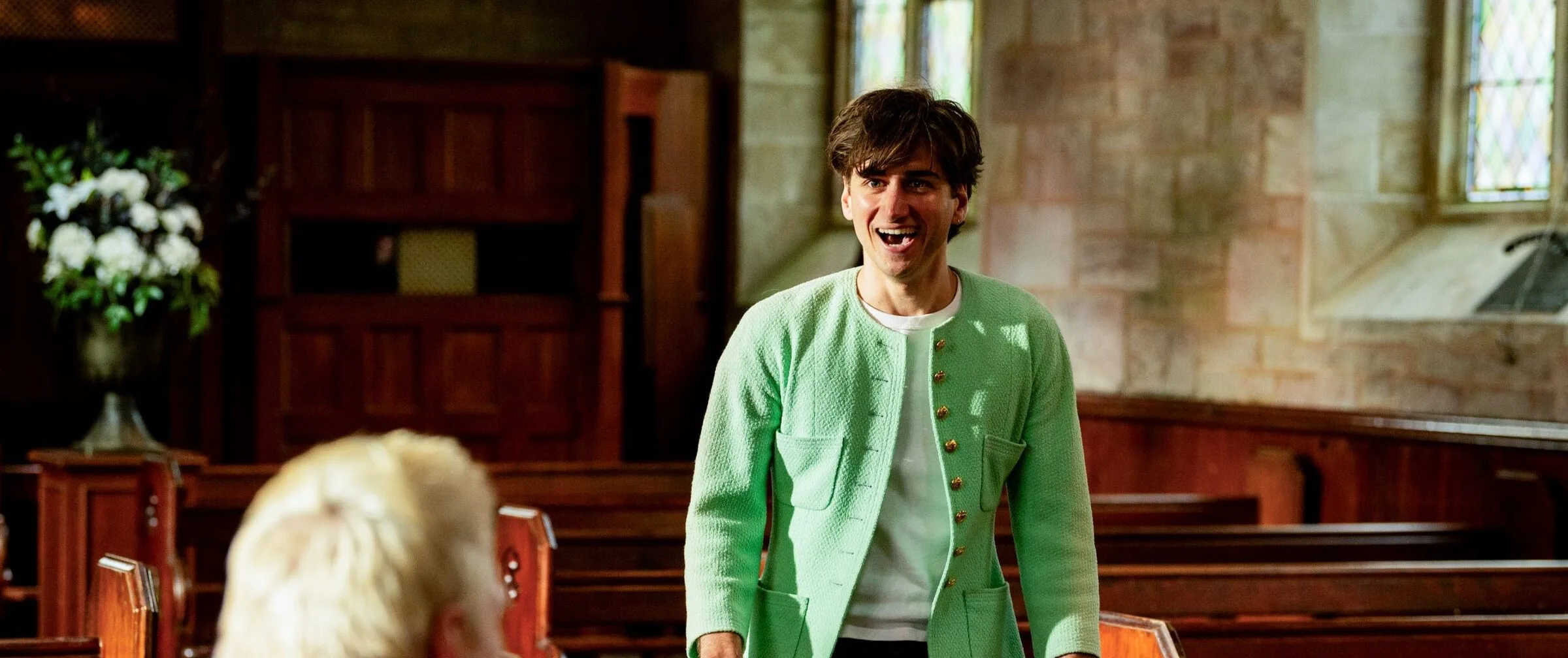Sebastian Croft laughing in the Celebrity Traitors chapel in a mint-green bouclé collarless jacket with gold buttons over a white T-shirt, with another contestant out of focus in the foreground.
