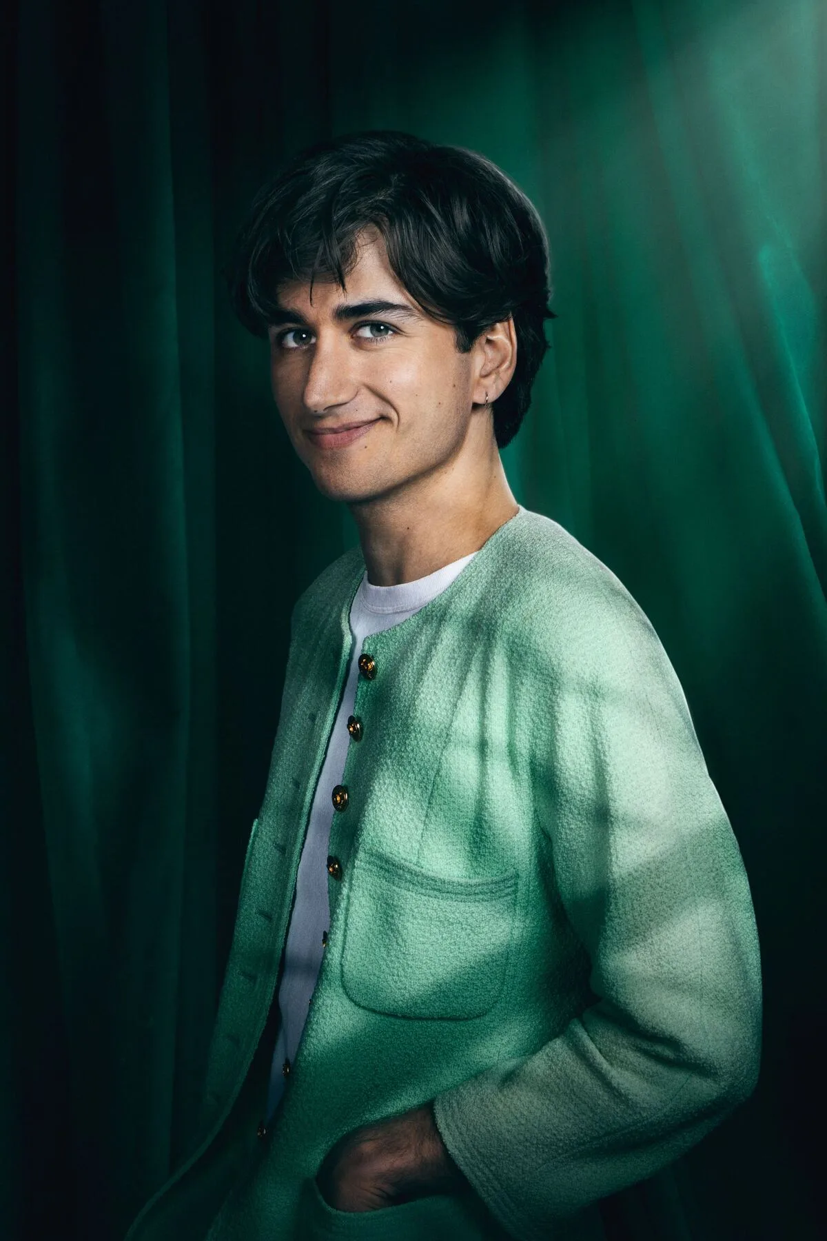 Sebastian Croft in a Celebrity Traitors cast portrait against green velvet curtains, wearing a mint-green textured collarless jacket with patch pockets and gold buttons over a white T-shirt.