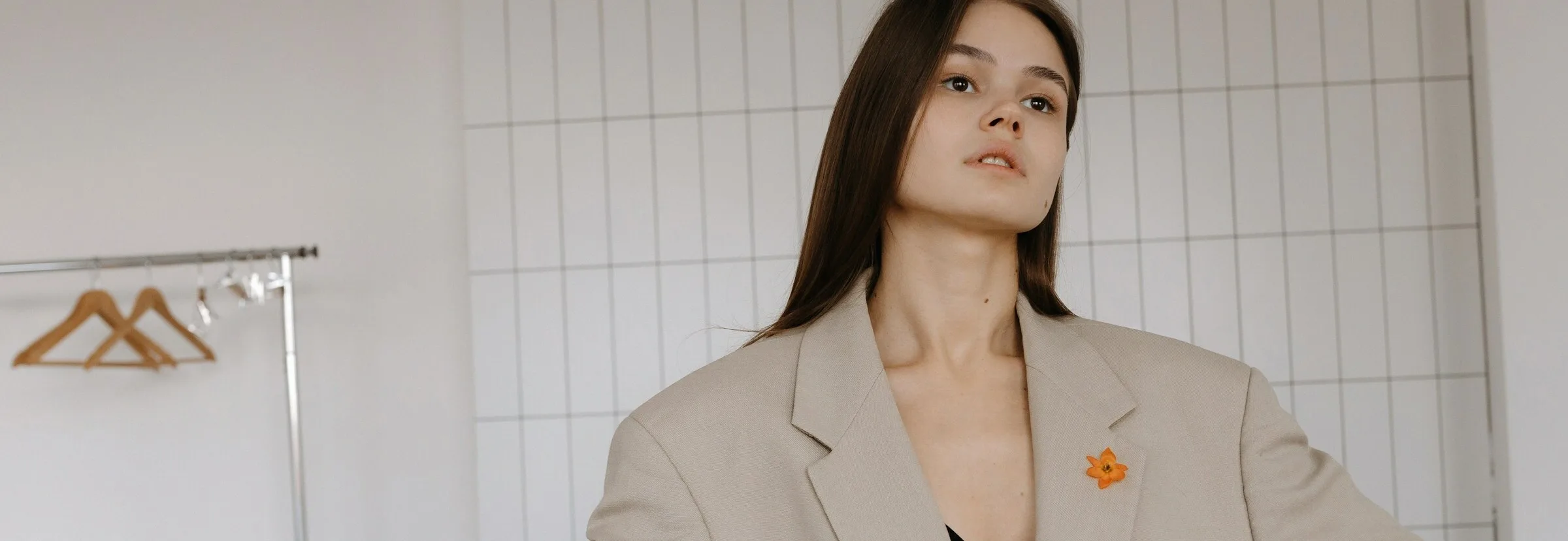 A woman wearing an oversized stone-coloured blazer over a black bra top, with a small orange flower pinned to the lapel, standing in a white tiled studio beside a clothes rail.
