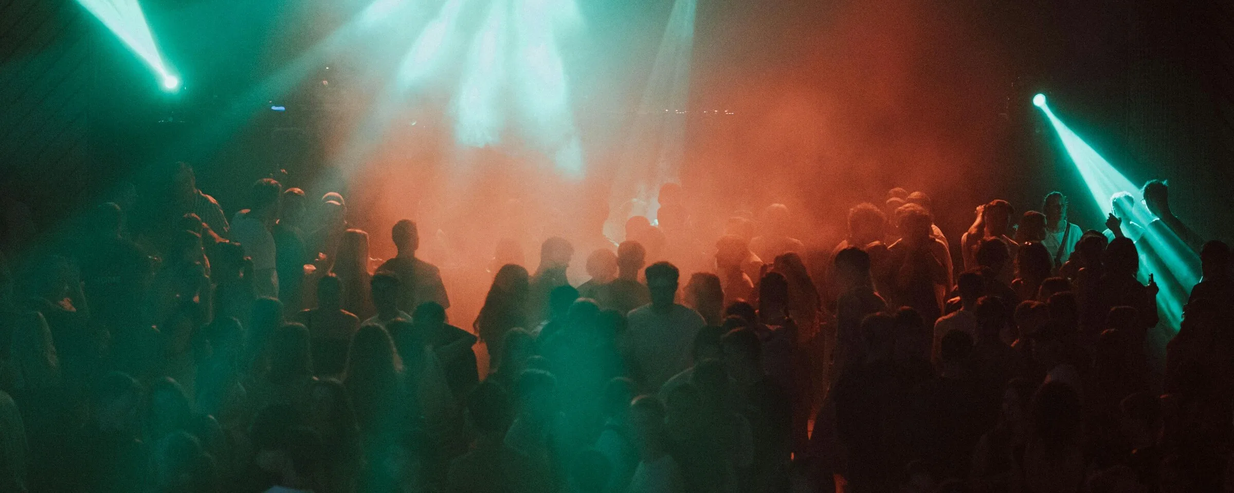 A packed indoor crowd at a warehouse gig, lit by teal and red stage beams through haze, with white tees and vest tops visible in the front rows.