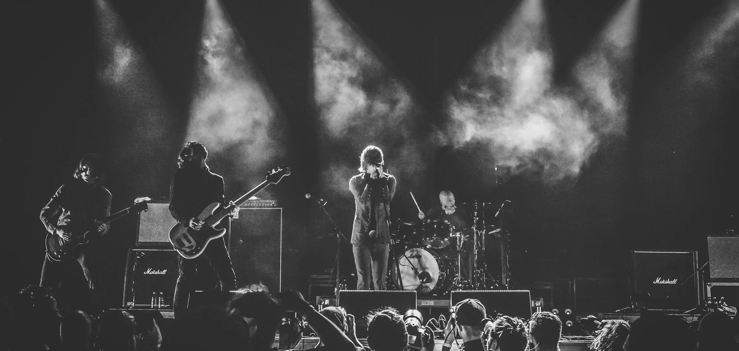 Black and white photo of a four-piece guitar band mid-set on a small club stage, lit from behind through haze, Marshall amps stacked either side