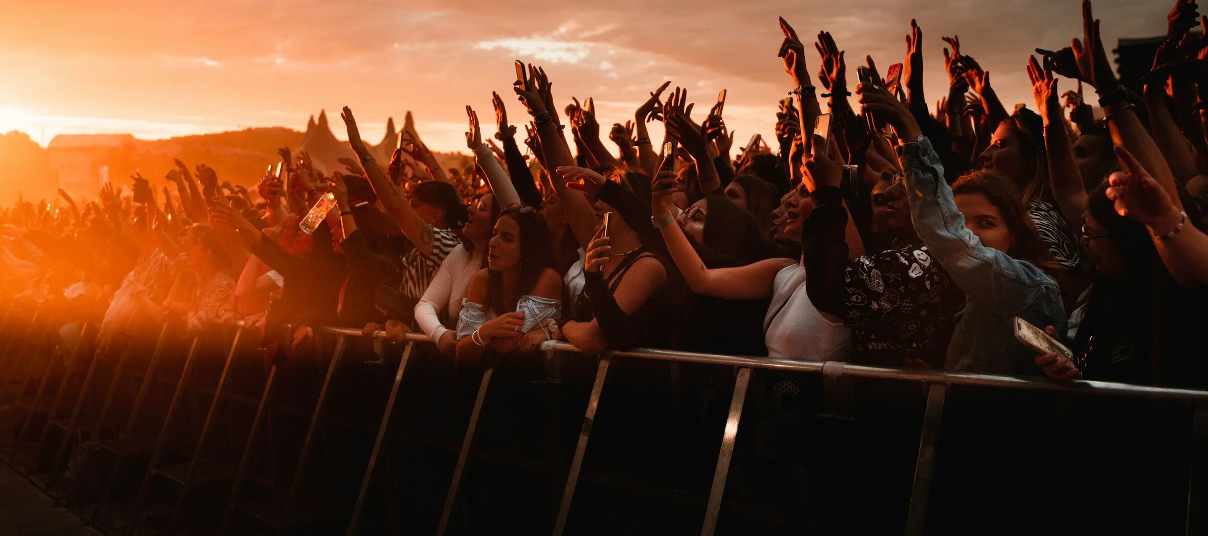 A packed crowd pressed against the front barrier at an outdoor gig at golden-hour sunset, hands and phones in the air, wearing bardot tops, black vests, a striped top and a denim jacket