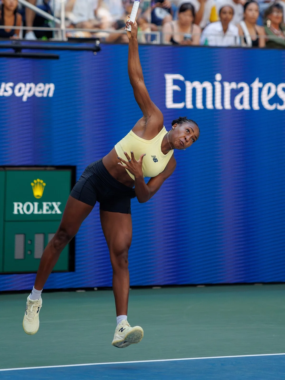 Coco Gauff mid-serve at the 2026 US Open in a pale butter yellow New Balance crop top, black shorts and pale yellow trainers