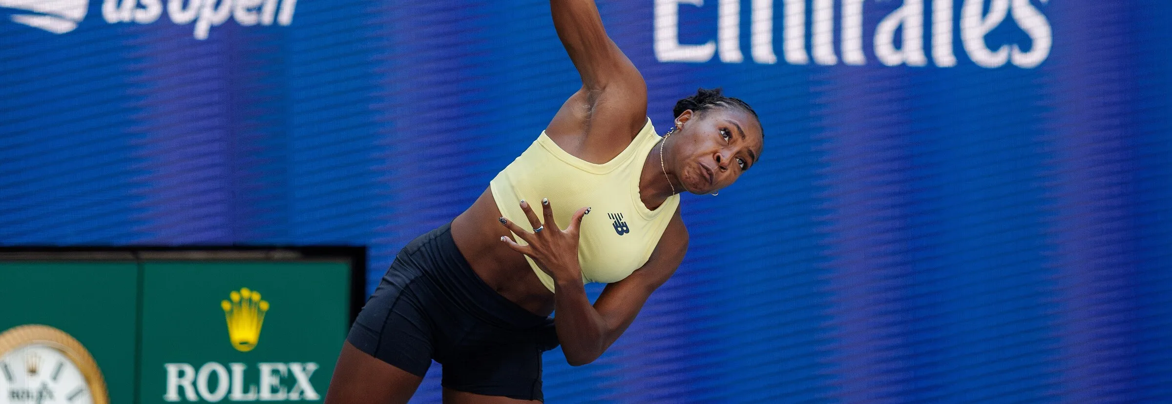 Coco Gauff serving at the 2026 US Open in a pale butter yellow New Balance crop top and black shorts, against a deep blue US Open backdrop