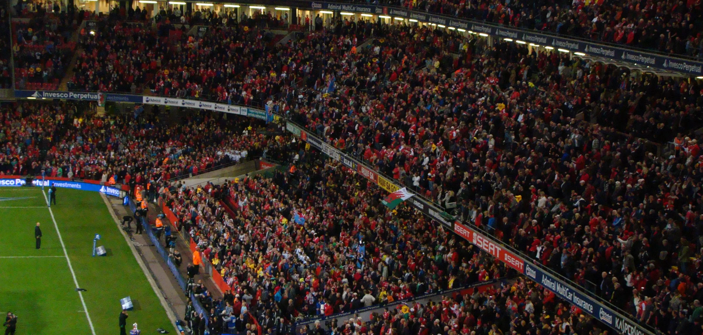 Wales supporters, most of them in red, fill the lower and upper tiers of the Principality Stadium under floodlights, with a Welsh dragon flag held up in the crowd and the edge of the pitch in the bottom left corner.